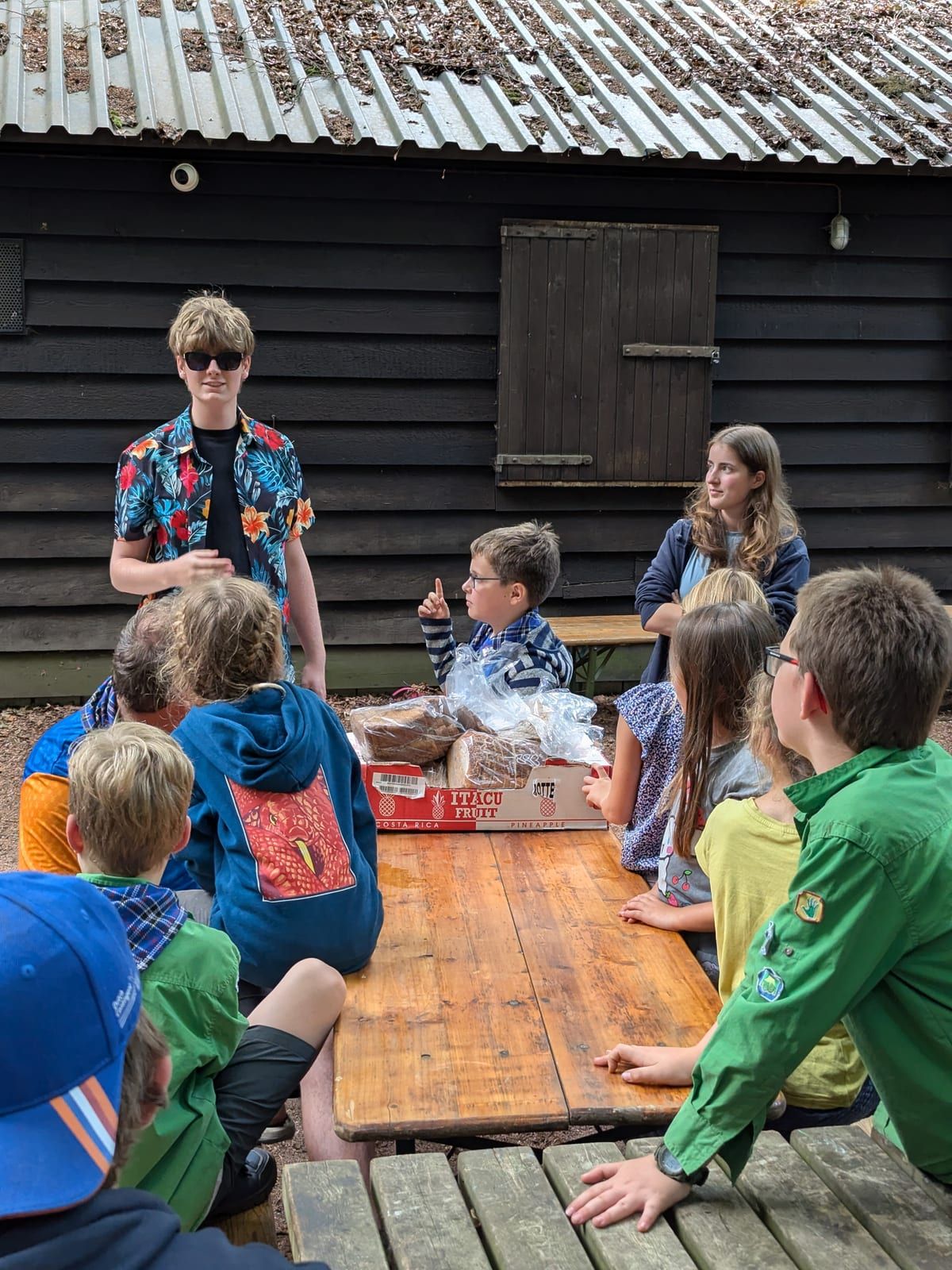 Welpen aan een picknicktafel bij de blokhut, met leiding erbij